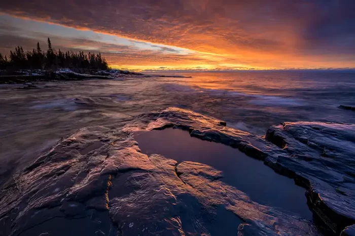 A wave pool next to Lake Superior surrounded by basalt with waves on the lake during a red sunrise.